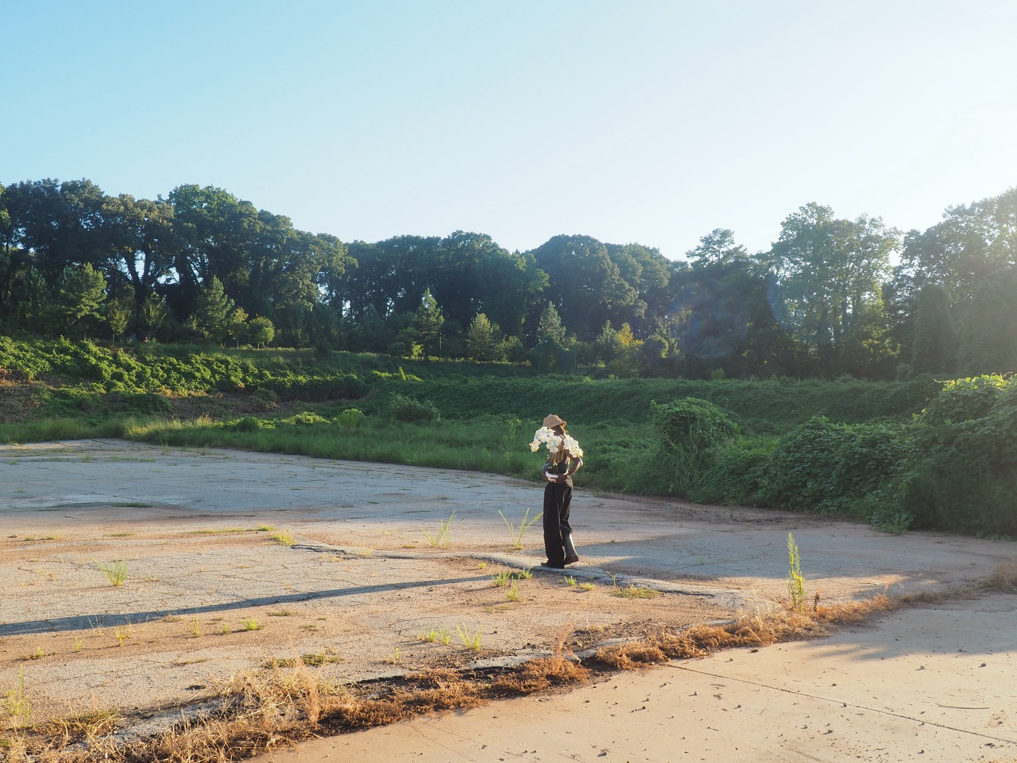 W.J. Lofton, small in the frame, stands on cracked pavement at the edge of a green field in low sun, holding white orchids.