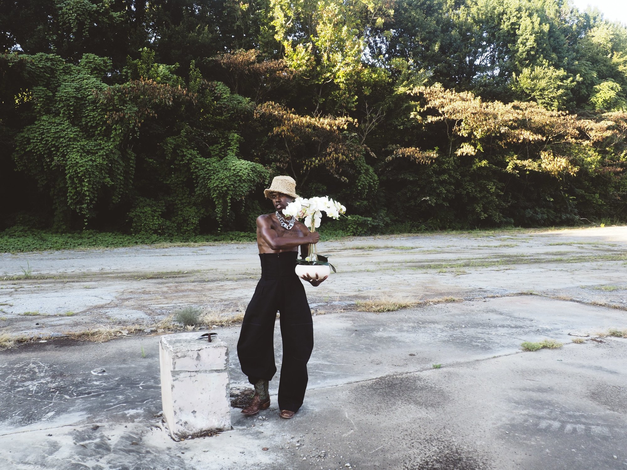 W.J. Lofton in a straw bucket hat, chain, and wide black trousers, cradling a bowl of white orchids in an overgrown lot beside a concrete block.