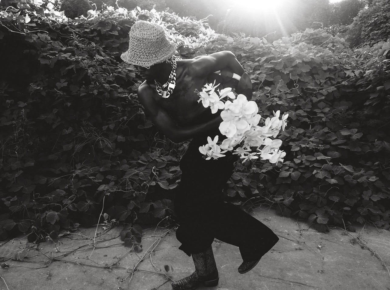 Black-and-white photograph of W.J. Lofton mid-stride before a wall of vines, in a woven bucket hat, heavy chain, and cowboy boots, carrying a spray of white orchids.